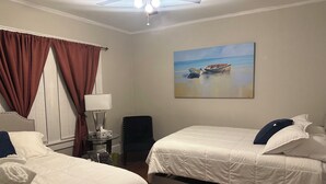 Habitación de hotel con dos camas, un ventilador de techo, un cuadro de un barco en la playa y una ventana con cortinas.