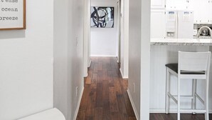 A hallway with wooden flooring, white walls, and a kitchen area with white cabinets and a bar stool.