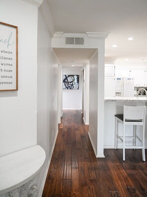 A hallway with wooden flooring, white walls, and a kitchen area with white cabinets and a bar stool.