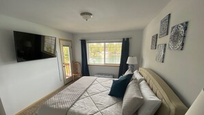 Une chambre à coucher avec un lit, un téléviseur fixé au mur, une fenêtre avec des rideaux et une porte donnant sur l’extérieur.