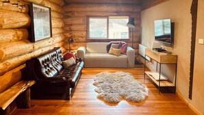 A log cabin interior with a leather sofa, a white couch, a fur rug, and a television on a wooden stand.