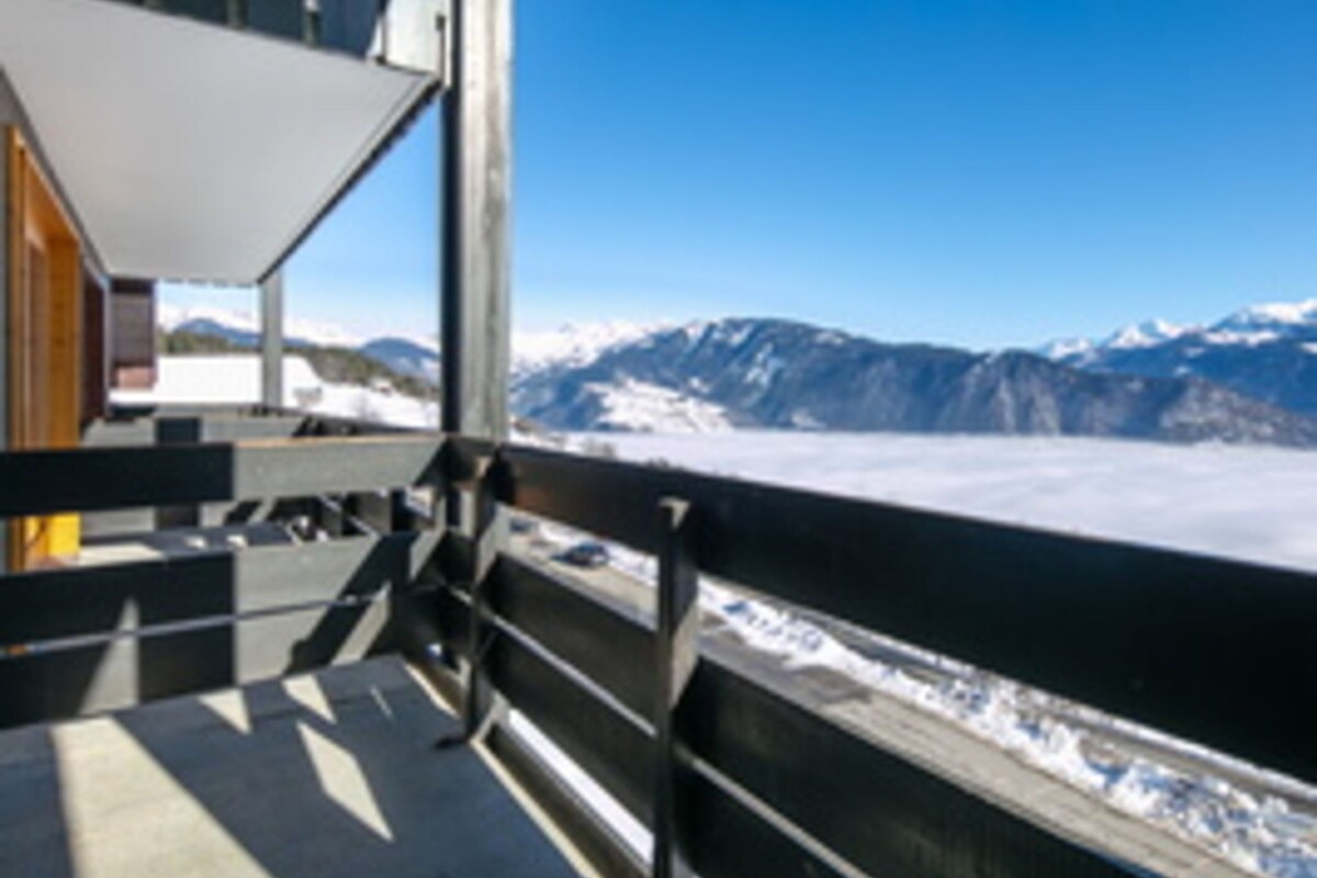A view from a window with a metal railing, overlooking a snow-covered landscape and mountains.