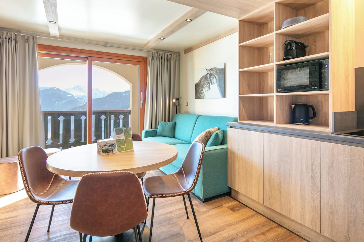 Habitación acogedora con una mesa redonda, dos sillas, un sofá azul verdoso, estantes de madera con electrodomésticos de cocina y vistas a las montañas desde un balcón.