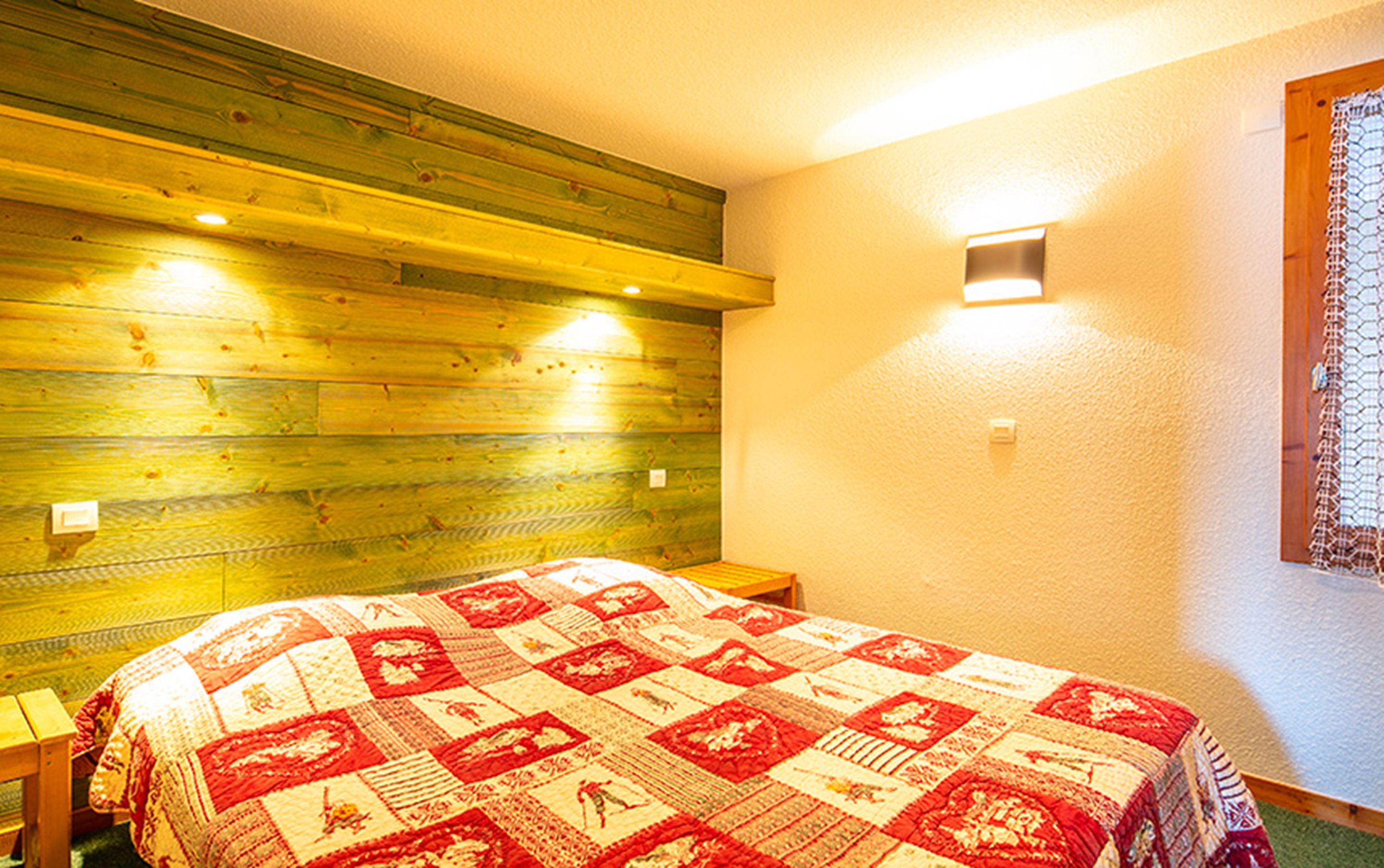 A bedroom with a wooden headboard, a quilted bedspread, a nightstand, and a wall-mounted light fixture.