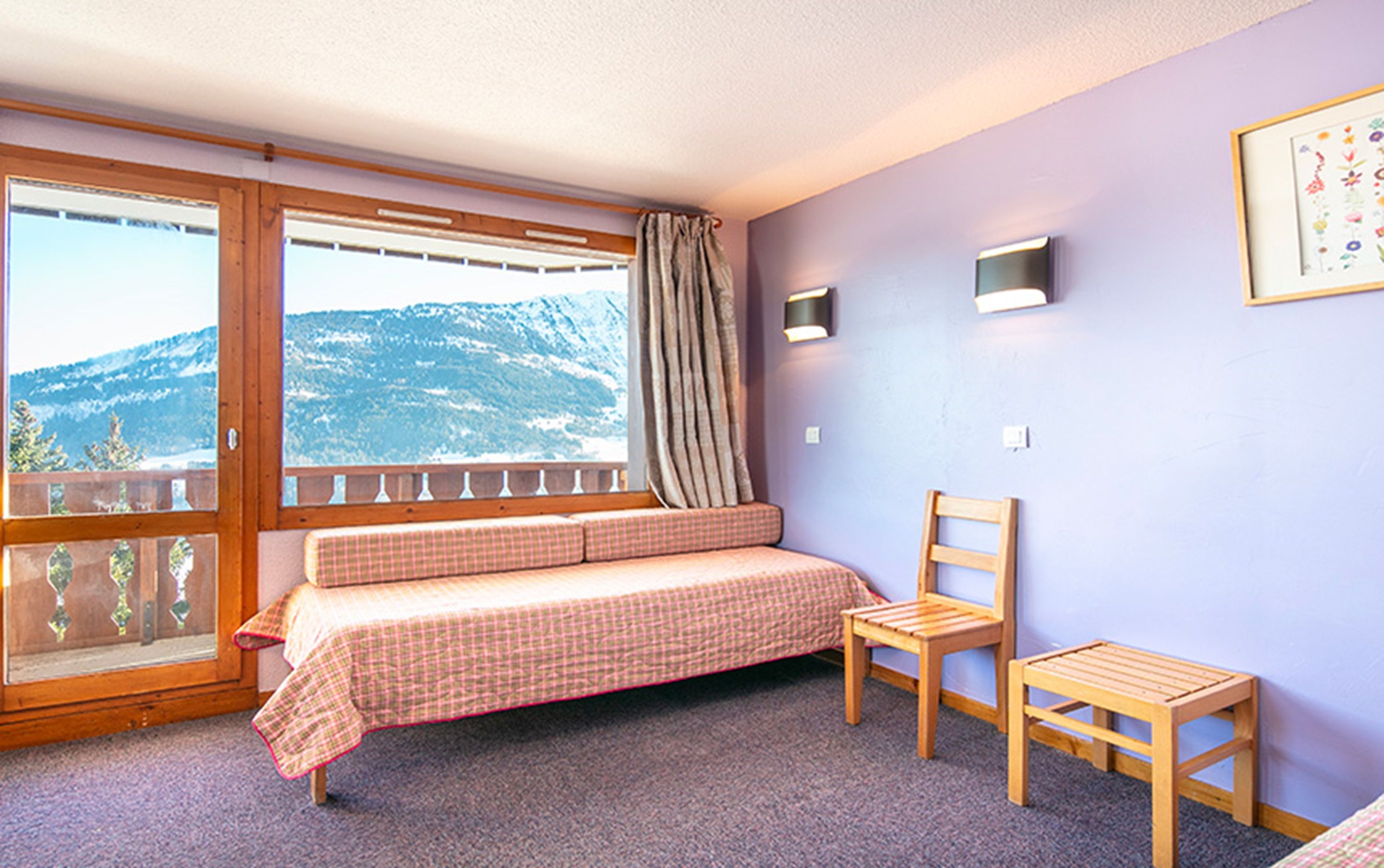 A room with a bed, a chair, a small table, a window with a view of mountains, and a wall-mounted light fixture.