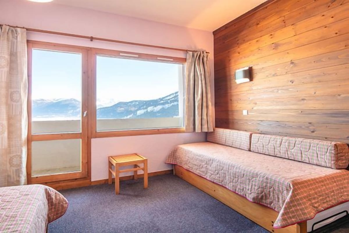 A cozy room with a wooden wall, a bed with a checkered blanket, a small wooden stool, and a window with a view of mountains.