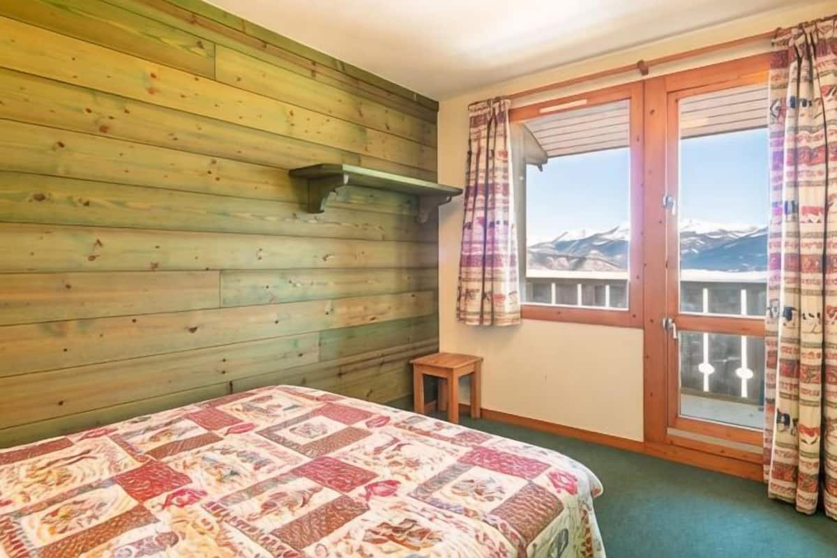 Un dormitorio con pared de madera, una cama con colcha estampada, un taburete de madera y una ventana con cortinas.