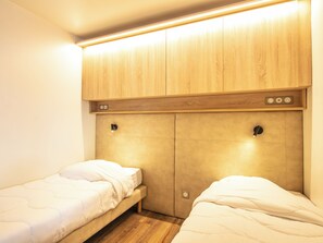 A bedroom with two single beds, wooden headboard, built-in lighting, and a small shelf with electrical outlets. - Residence Le Beauregard - Vacanceole (La Léchère)