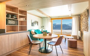 A cozy living area with a wooden floor, a round dining table with chairs, a green sofa, and a view of mountains through a large window. - Residence Le Beauregard - Vacanceole (La Léchère)