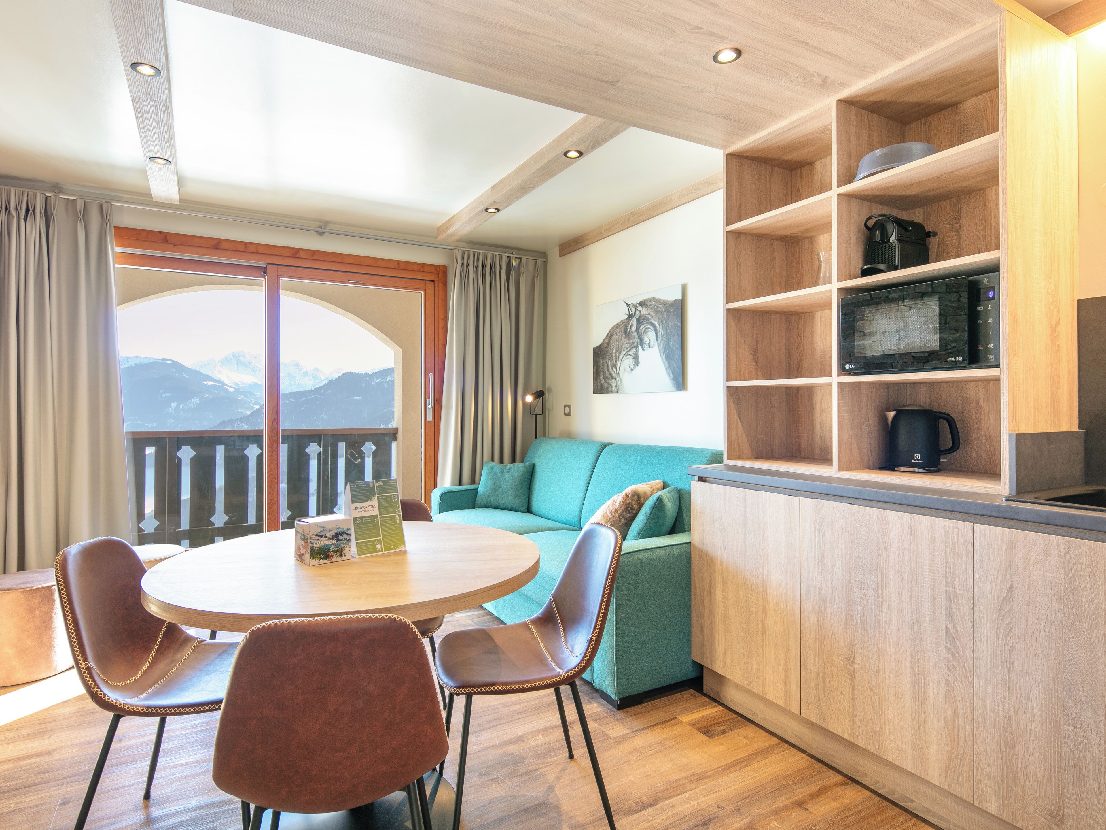 Habitación acogedora con una mesa redonda, dos sillas, un sofá azul verdoso, estantes de madera con electrodomésticos de cocina y vistas a las montañas desde un balcón.
