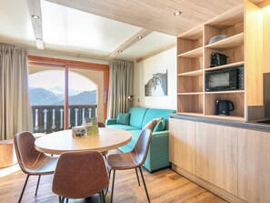 A cozy room with a round table, two chairs, a teal sofa, wooden shelves with kitchen appliances, and a view of mountains through a balcony. - Residence Le Beauregard - Vacanceole (La Léchère)