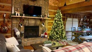 Interior acolhedor de cabana com lareira de pedra, árvore de Natal, sofás de couro e área de jantar.