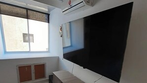 A room with a large flat-screen TV mounted on the wall, a wooden cabinet below it, a window with blinds, and an air conditioning unit on the wall. - Remodeled Gorgeous Apartment on Beach (Cartagena)