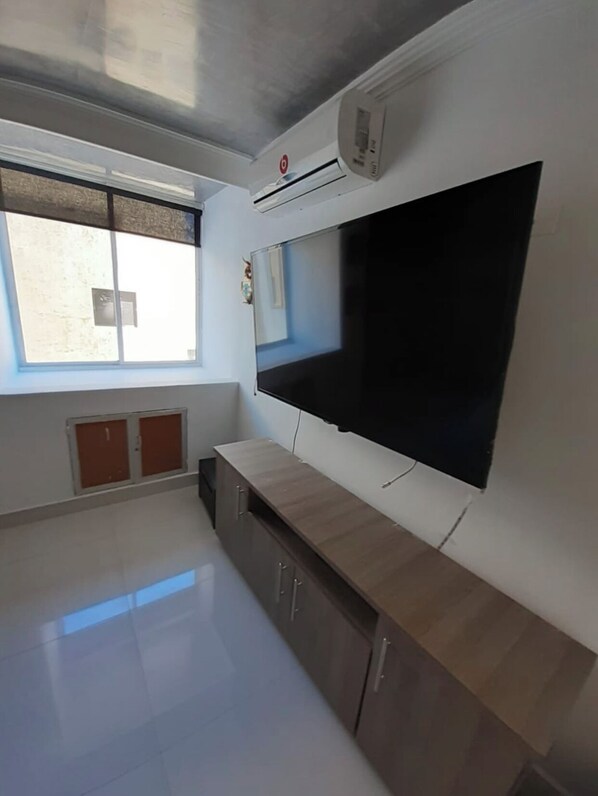 A room with a large flat-screen TV mounted on the wall, a wooden cabinet below it, a window with blinds, and an air conditioning unit on the wall.