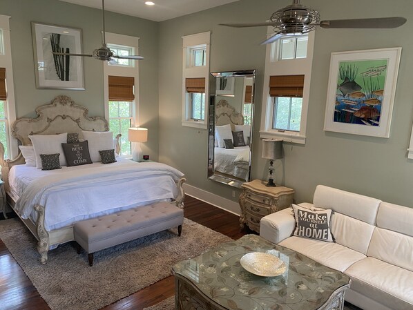 A bedroom with a large bed, a sitting area with a sofa, a glass coffee table, and a ceiling fan.