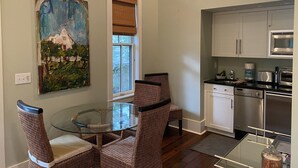 Een eetgedeelte met een glazen tafel en rieten stoelen, een keuken met witte kasten en moderne apparatuur, en een schilderij aan de muur.