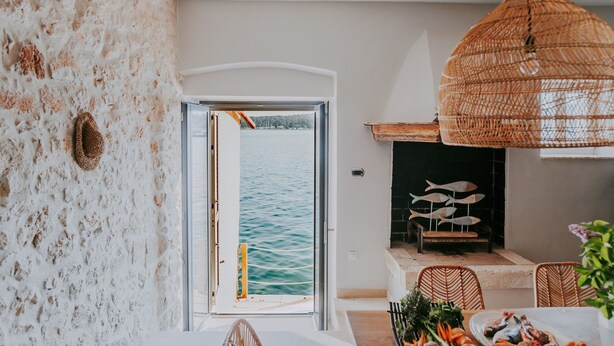 Ein Zimmer mit Steinwand, Meerblick durch eine offene Tür, Korbmöbel und ein Kamin mit Fischverzierungen.