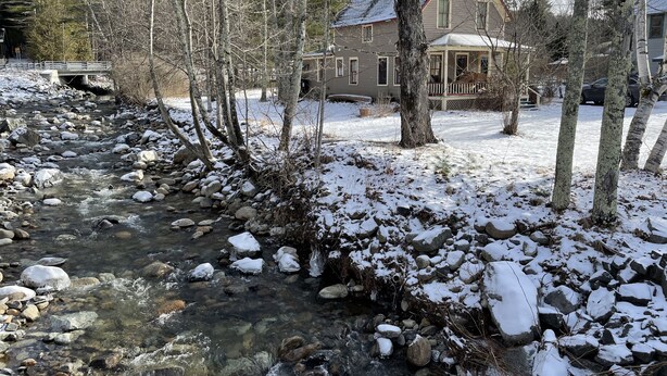 Un quartier résidentiel traversé par un ruisseau, au milieu d’un paysage enneigé, bordé d’arbres et de maisons.