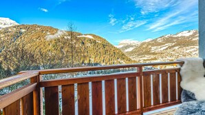 Holzgeländer eines Balkons mit Blick auf eine Berglandschaft, schneebedeckte Gipfel und einen klaren, blauen Himmel.