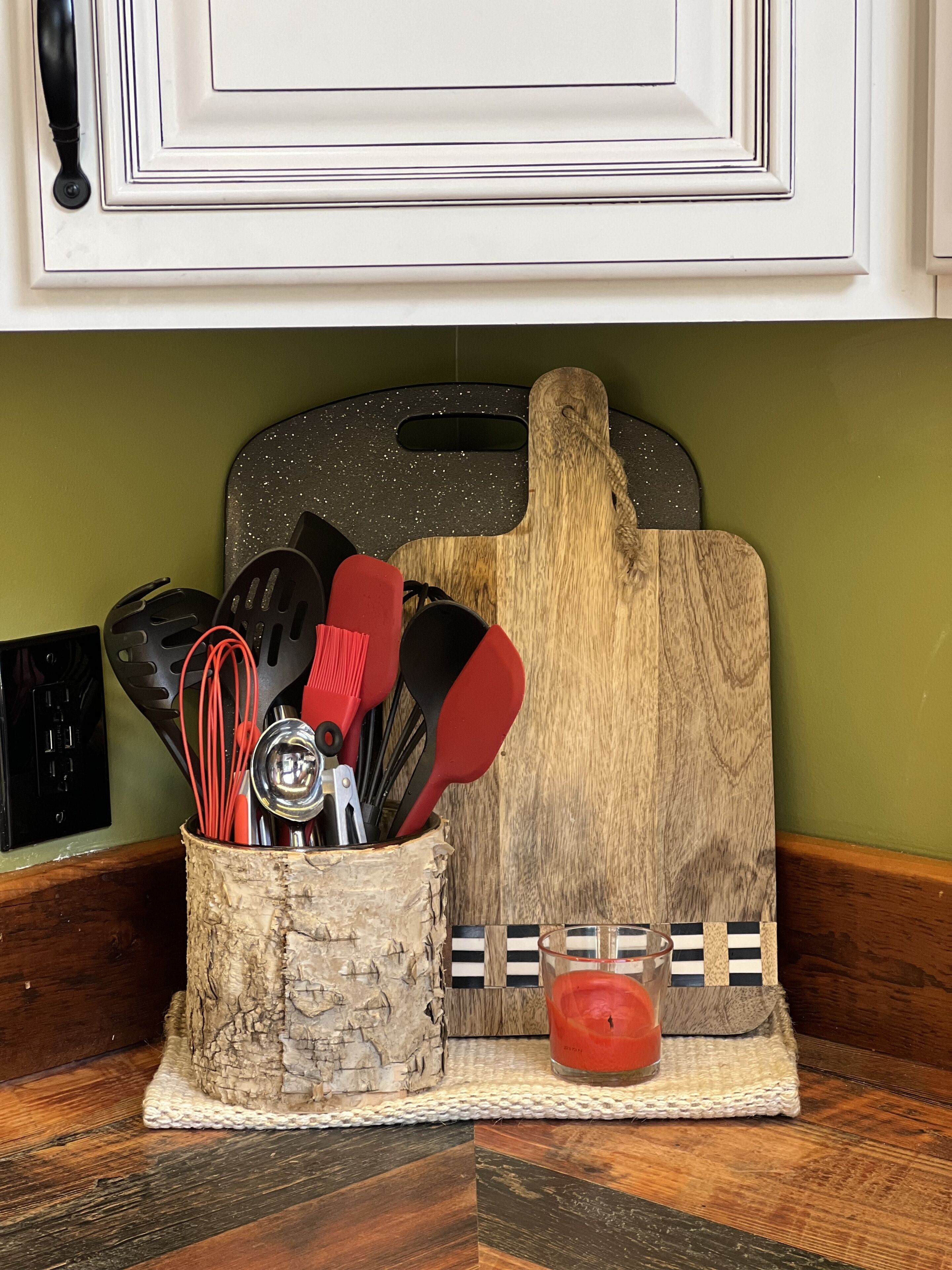 A kitchen counter with a wooden cutting board, a woven basket holding kitchen utensils, and a candle.