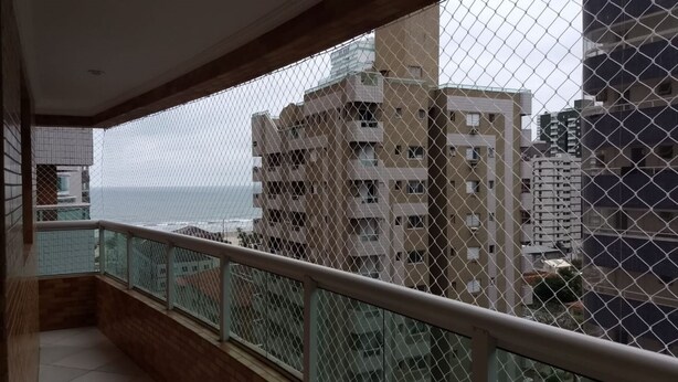 A balcony with a chain-link fence, overlooking a view of high-rise buildings and the sea. - New apartment in Canto do Forte with ocean view, close to the beach. (PRAIA GRANDE)