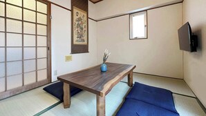 A traditional Japanese room with tatami flooring, a low wooden table, a blue cushion, a wall-mounted TV, and a shoji screen door.