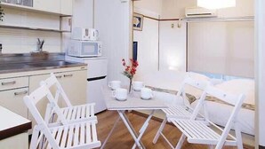 Cocina compacta con armarios blancos, microondas y un refrigerador pequeño. Hay una zona de comedor con sillas blancas y una mesa, además de un ventanal con vistas.