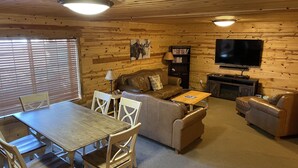 Un interior acogedor de cabaña con piso de madera, una mesa de comedor con sillas, un sofá, un televisor y una estantería.