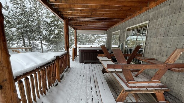 Un área de estar al aire libre techada, con bancos de madera y un jacuzzi, rodeada de árboles cubiertos de nieve.