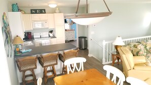 A kitchen with white cabinets, a microwave, and a stove. There is a dining area with a wooden table and wicker chairs. - Surf City • Views, Views, and more Views! (Surf City)