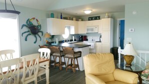 A compact kitchen with white cabinets, a central island with bar stools, a dining area with a white table and chairs, and a yellow armchair. - Surf City • Views, Views, and more Views! (Surf City)