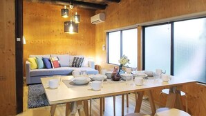 Un intérieur chaleureux en bois, avec un coin repas comprenant une longue table et six tabourets, un canapé agrémenté de coussins colorés et une grande fenêtre laissant entrer la lumière naturelle.