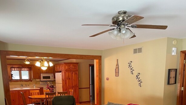 A living room with a brown leather sofa, a wooden coffee table, and a patterned rug. There is a dining area with wooden cabinets and a chandelier. A ceiling fan is visible above the living room. - Quiet Lake with Kayaks and Direct Beach Access! (Neshkoro)