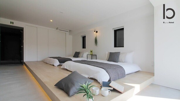 A modern hotel room with two beds, a small bedside table, a plant, and a wall-mounted light fixture. - b hotel 1002 Exclusive Urban Penthouse Stay (Hiroshima City)