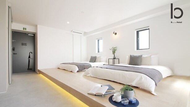 A modern hotel room with a minimalist design, featuring two beds, a small seating area with a book and a plant, and a visible bathroom door.