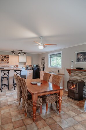 A modern kitchen with a dining area, featuring a large wooden table, bar stools, a built-in stove, and a stone fireplace. - Omas retreat...Romantic Getaway (Blampied)