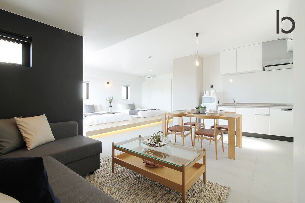 A modern hotel room with a dark accent wall, a sofa, a glass coffee table, a dining area with a wooden table and chairs, and a kitchenette.