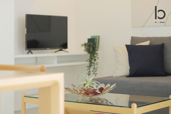 Una sala de estar moderna con un televisor de pantalla plana montado en una pared blanca, una mesa de centro de vidrio con una planta decorativa y un sofá gris con un cojín oscuro.