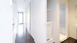 Un couloir moderne et bien éclairé, doté d’un sol carrelé foncé, qui mène à une salle de bain avec un lavabo blanc et un espace douche.