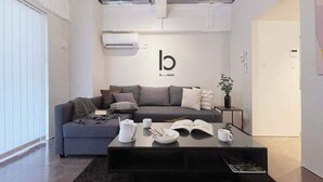 Une chambre d’hôtel moderne avec un canapé gris, une table basse noire et un mur blanc orné d’un logo.