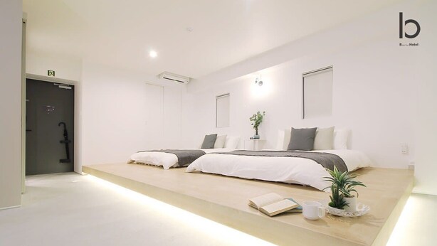 Une chambre d’hôtel moderne au design minimaliste, comprenant deux lits, une table de chevet avec un livre et une tasse, et une petite plante en pot.