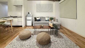 Una sala de estar moderna con un sofá gris, una mesa de centro, dos otomanas redondas, una pequeña cocina con microondas y una pared grande y despejada.