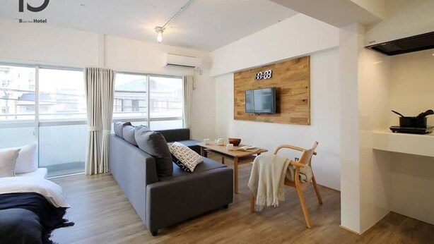 Una habitación de hotel moderna con un sofá, una pequeña mesa de comedor, un televisor montado en una pared de madera y un gran ventanal con cortinas.