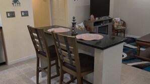 Un coin repas comprenant une table avec un plateau en verre, deux chaises en bois, un tapis décoratif et un téléviseur posé sur un meuble.