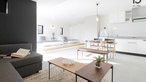 Een moderne hotelkamer met een donkergrijze bank, een houten salontafel en een keukenhoek met witte kasten en een magnetron.
