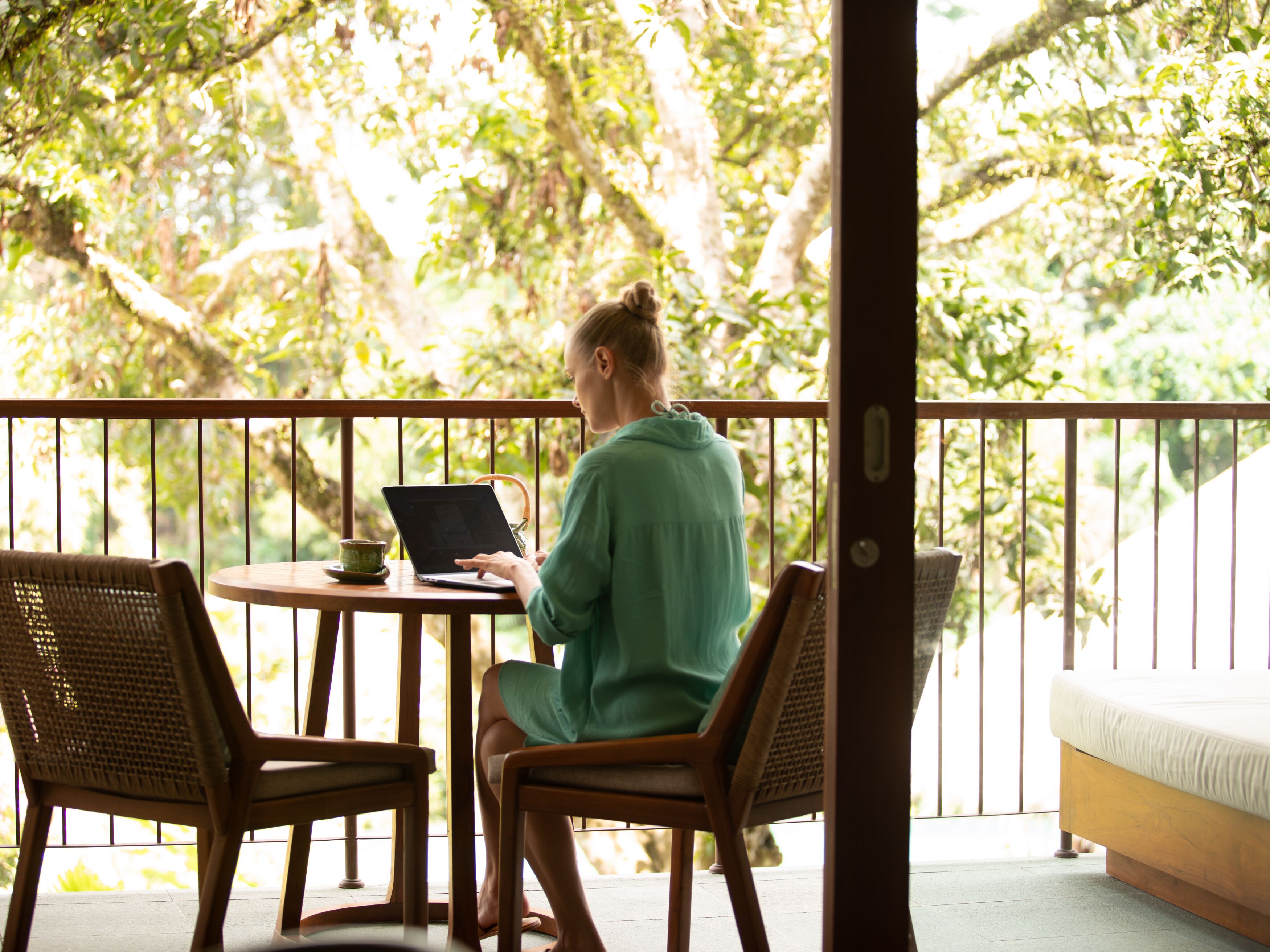 Une personne est assise à une table, un ordinateur portable posé devant elle, sur un balcon agrémenté de chaises en bois et d’une petite table, le tout entouré de verdure.