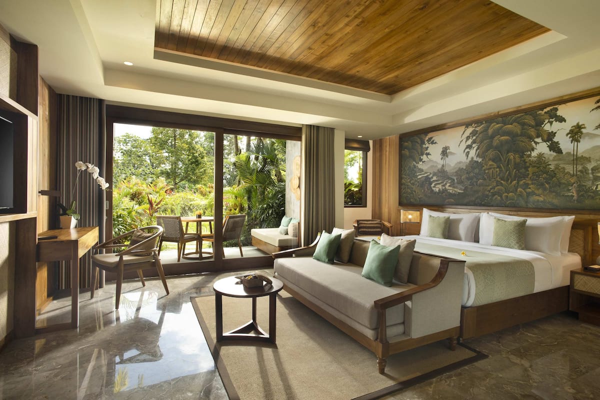 A modern hotel room with a large bed, a sofa, a dining area, and a view of lush greenery.