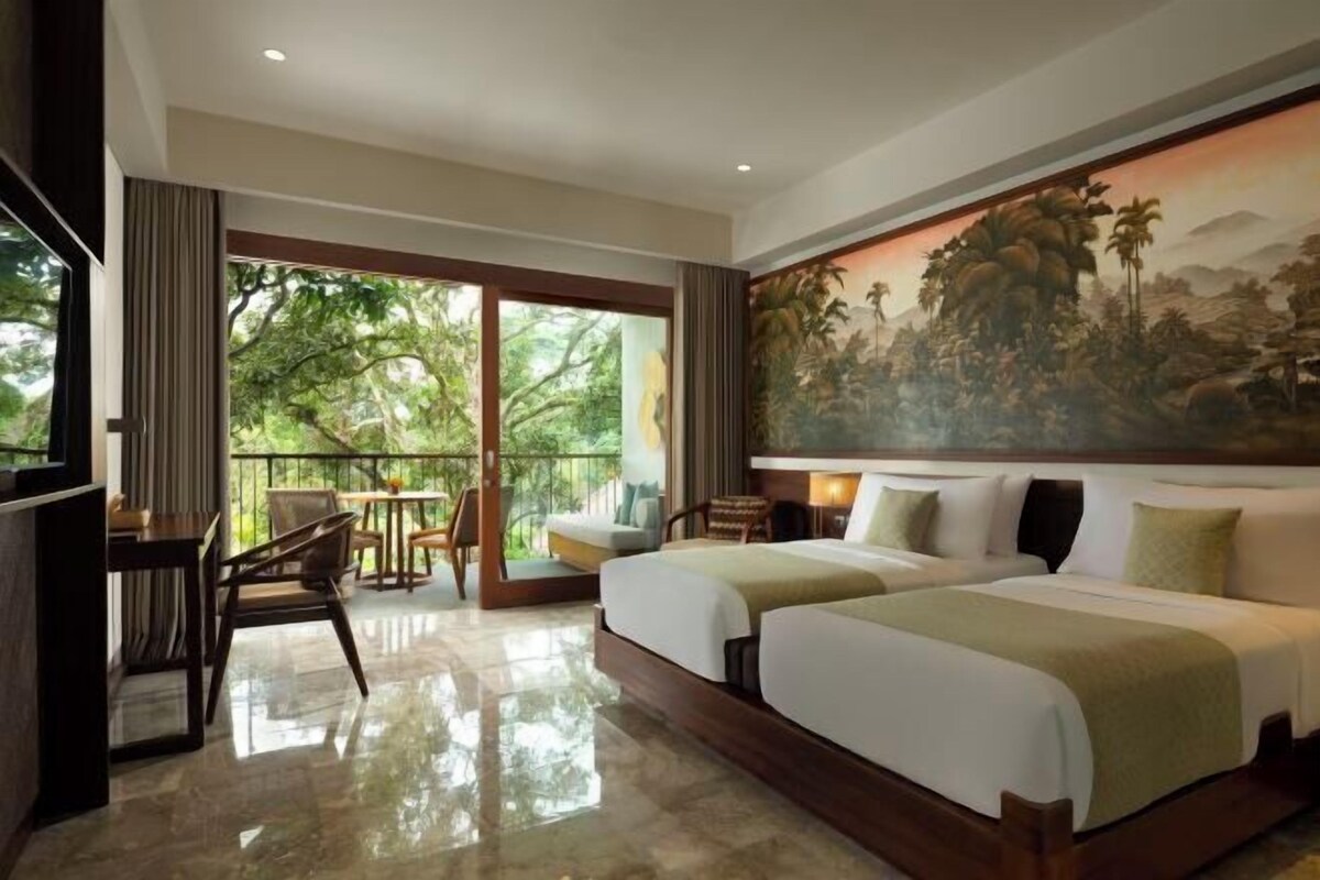 A hotel room with two beds, a balcony with a view of greenery, a painting on the wall, and a dining area with chairs and a table.
