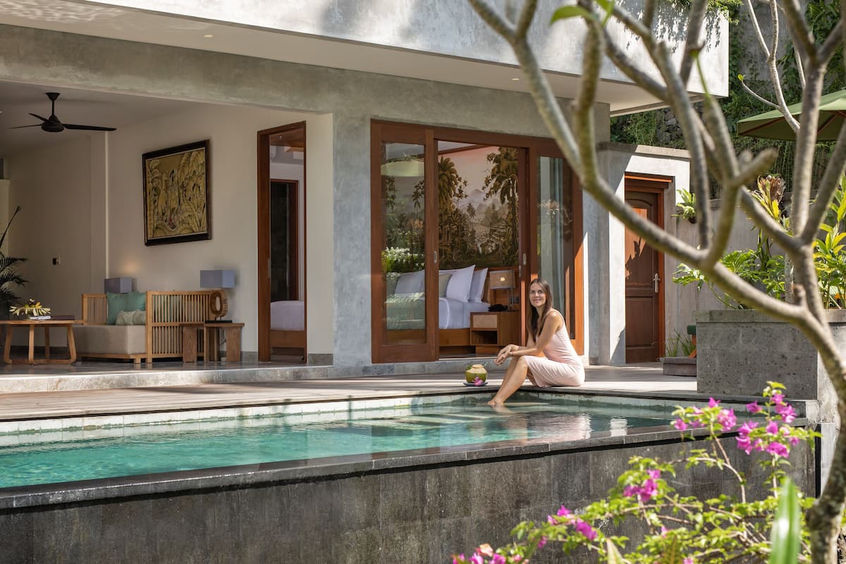 A modern pool area with a person sitting by the pool, a wooden deck, and a room with a bed and a window.