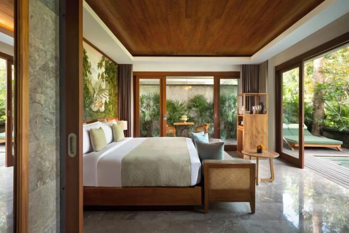 A modern bedroom with a large bed, a small table, and a chair. There is a view of a garden through large windows.